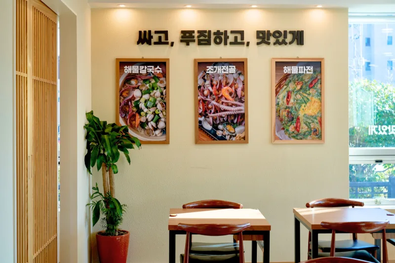 울산 식당인테리어 시공사례 — 해운대31해물칼국수 동구점 울산 동구, 메뉴 액자를 건 밝은 크림색 벽과 원목 테이블